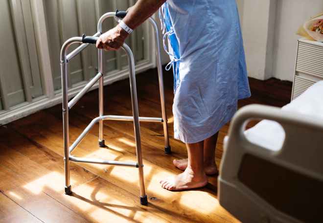 person in hospital gown using walking frame beside hospital bed
